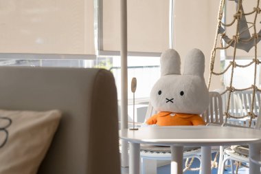 Büyük peluş, turuncu elbiseli tavşan oyuncağı. Kapalı bir mekanda güneşli bir pencerenin yanında, sandalyede oturuyor.