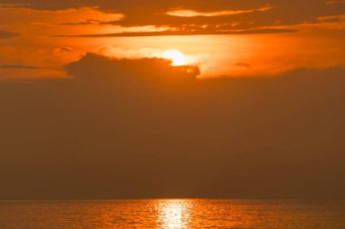 Okyanusun üzerinde dramatik turuncu gün batımı. Güneş kısmen kara bulutlarla kaplı ve sakin su yüzeyine yansıyor.