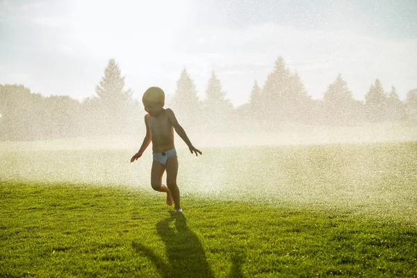 Маленький мальчик бежит под фонтанами в зеленых полях — стоковое фото