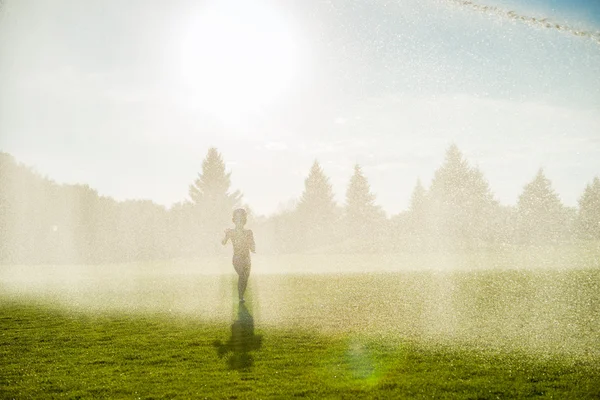 Un petit garçon court sous les fontaines dans les champs verts — Photo