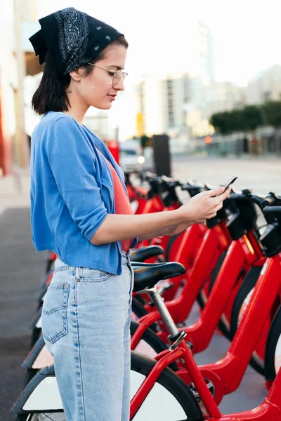 vertical photo of a young woman using the phone to unlock a bike from the rental station, concept of ecology and sustainable mobility against climate change, copy space for text