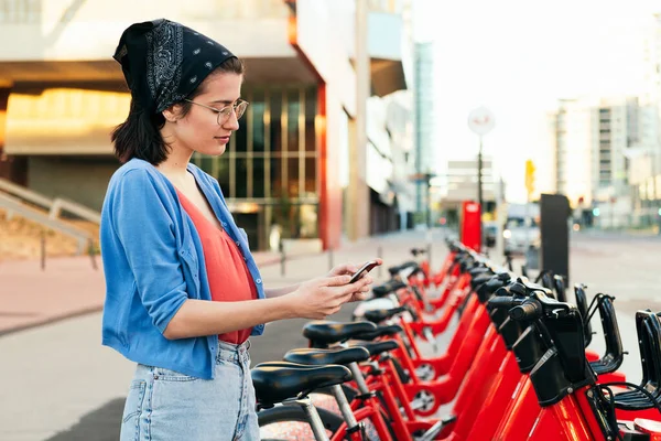 young woman using a smart phone next to an electric bike rental station in the city, concept of ecology and sustainable mobility against climate change, copy space for text