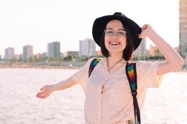 Şehrin yanındaki deniz kenarında mutlu bir şekilde gülümseyen şapkalı genç bir gezginin portresi, seyahat ve turizm konsepti, metin için kopyalama alanı