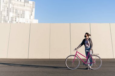 Koruyucu maskeli genç bir kadın şehirde pembe bir retro motorla duruyor, aktif yaşam tarzı kavramı, covid ve sürdürülebilir kentsel hareketliliğe karşı koruma