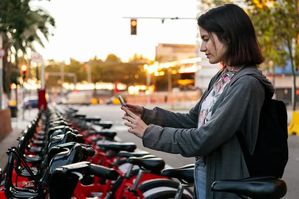young girl taking a bike with her smart phone in a public parking of rental bicycles in the city, concept of active lifestyle and sustainable mobility