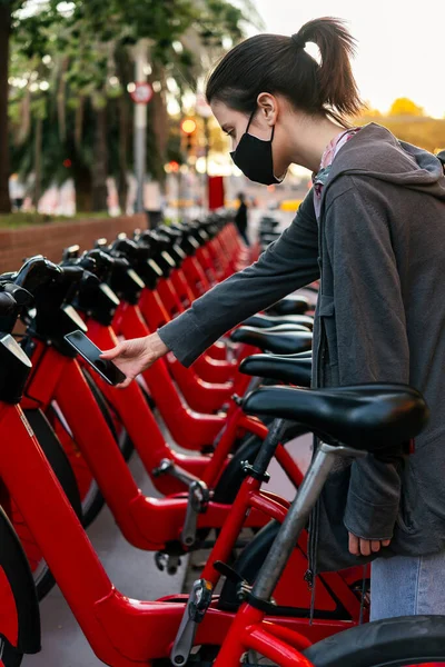 vertical photo of a young girl with protective mask reading the code with a smart phone to unlock a bicycle from the urban bike rental parking, concept of active lifestyle and sustainable mobility