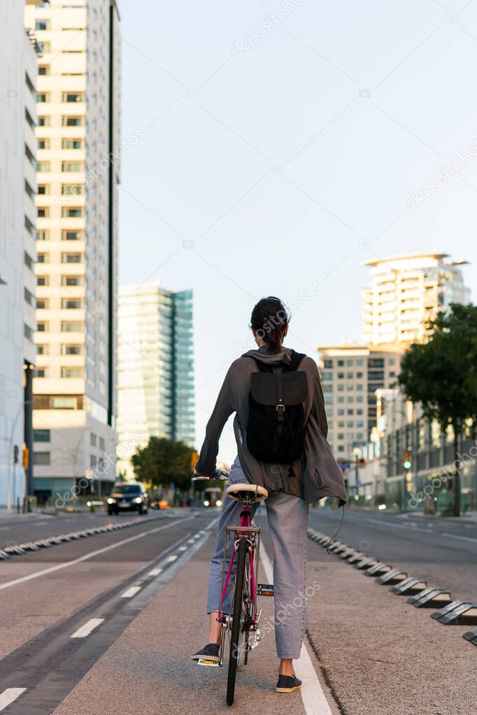 vertical rear view of a young girl with backpack at the city on a pink retro bike, concept of active lifestyle and sustainable mobility