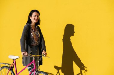 Renkli sarı bir duvarın önünde yürüyen gülümseyen genç bir kadın pembe retro bisikleti, aktif yaşam tarzı ve sürdürülebilir hareketlilik kavramı, metin için kopyalama alanı
