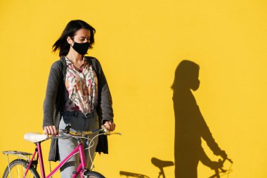 young girl wearing protective mask walks with her vintage pink bike in front of a colorful yellow wall, concept of protection against covid and sustainable mobility, copy space for text