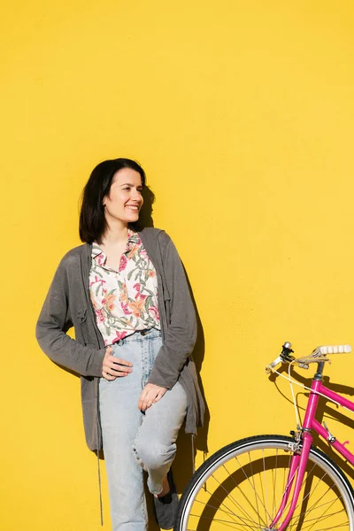 vertical portrait of a happy young woman leaning against a colorful yellow wall next to her pink retro bike, concept of active lifestyle and sustainable mobility, copy space for text