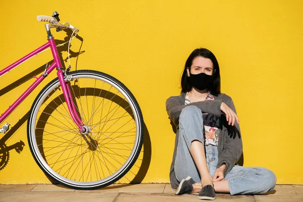 young girl wearing protective mask sitting next to her pink retro bike leaning on a colorful yellow wall, concept of protection against covid and sustainable mobility, copy space for text