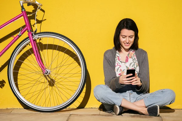 smiling young woman consulting smart phone sitting next to her bike leaning against a colorful yellow wall, concept of technology and sustainable mobility