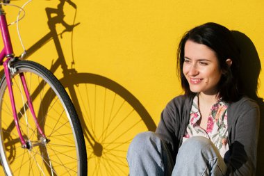 portrait of a happy young girl sitting next to her retro pink bike leaning against a colorful yellow wall, concept of active lifestyle and sustainable mobility