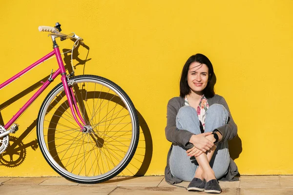 happy young girl sitting next to her retro pink bike leaning against a colorful yellow wall, concept of active lifestyle and sustainable mobility