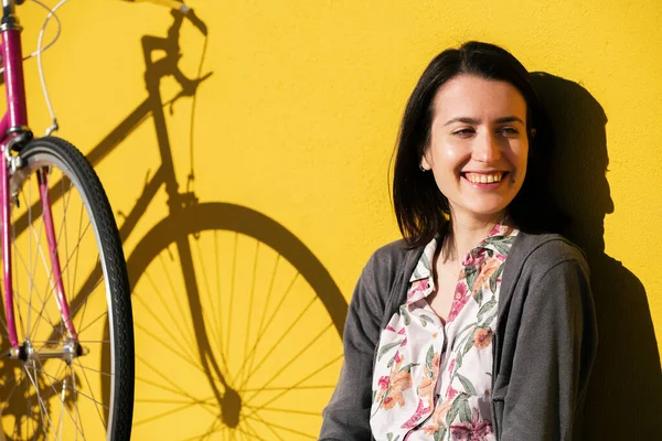 portrait of a smiling young girl sitting next to her retro pink bike leaning against a colorful yellow wall, concept of active lifestyle and sustainable mobility