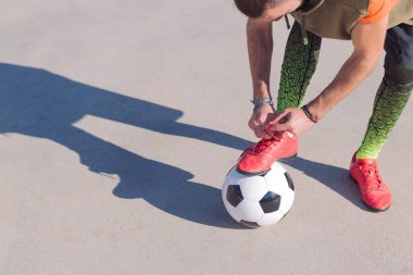 Ayağı beton bir futbol sahasında botlarını bağlayan tanınmamış bir futbolcu. Şehirde sağlıklı yaşam tarzı ve şehir sporu kavramı. Metinler için kopya alanı.