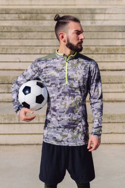 Beton bir futbol sahasında kolunun altında futbol topu olan çekici bir hippi sporcunun dikey fotoğrafı. Şehirde sağlıklı yaşam tarzı ve şehir sporu kavramı.