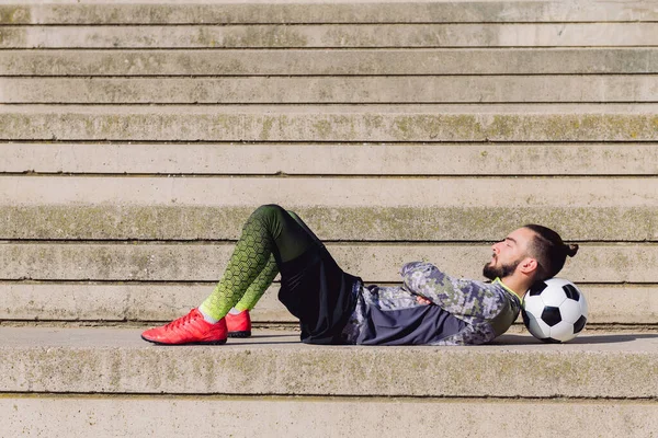 handsome sportsman with hair in a bun lying in a concrete soccer court with head on his football ball, concept of healthy lifestyle and urban sport in the city, copy space for text