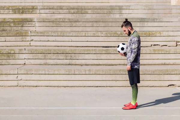 handsome sportsman with hair in a bun standing with the ball in his hand on a concrete soccer court, concept of healthy lifestyle and urban sport in the city, copy space for text