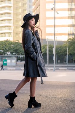 Şehir merkezinde şapkalı ve trençkotlu genç bir Asyalı kadının dikey fotoğrafı.