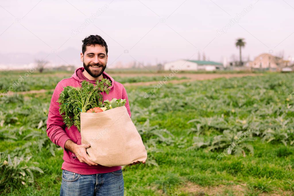 young entrepreneur farmer smiling with a burlap bag full of vegetables and fruits, concept of entrepreneurship in the countryside and ecological farming, copy space for text