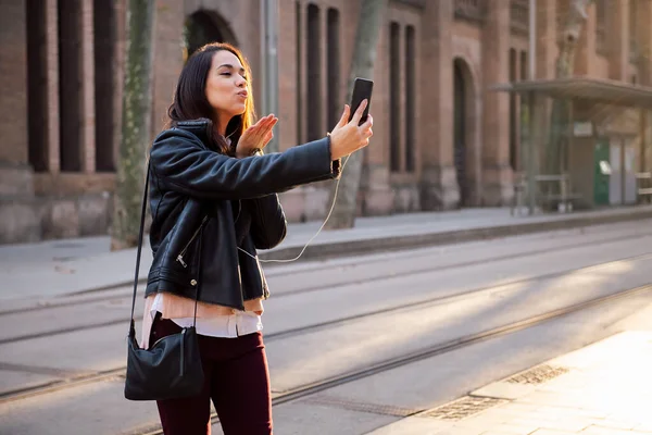 young woman sending a kiss by making a video call with her phone from the street, technology and communication concept, copy space for text