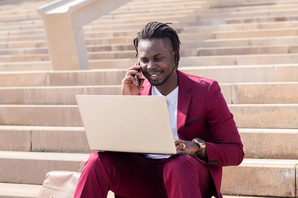 smiling black man works with computer laptop and phone sitting on city stairs, technology and remote work concept