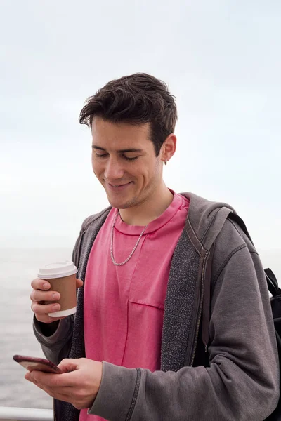 Bir fincan kahveyle gülümseyen ve cep telefonu kullanan mutlu bir gencin dikey fotoğrafı. Teknoloji ve şehir yaşam tarzı kavramı.