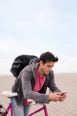 Sırt çantasıyla bisiklete yaslanan yakışıklı bir gencin dikey fotoğrafı cep telefonu, iletişim konsepti ve ekolojik şehir ulaşımı, metin için kopyalama alanı
