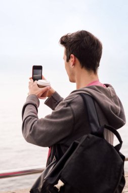 Sırt çantası ve kahveyle fotoğraf çeken genç bir öğrencinin arka planı cep telefonu, teknoloji konsepti ve milenyum yaşam tarzı, metin için kopya alanı, dikey fotoğraf