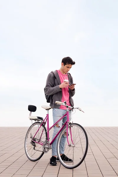 Pionowe Zdjęcie Młodego Człowieka Rowerem Trzymającym Kawę Podczas Korzystania Telefonu — Zdjęcie stockowe