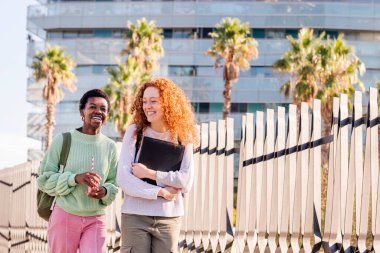 Sırt çantası ve klasörle kaldırımda yürürken mutlu gülümseyen iki genç kadın, öğrenci ve şehir gençliği yaşam tarzı kavramı, metin için kopyalama alanı