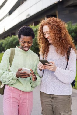 Mutlu gülümseyen ve cep telefonlarına bakan iki genç kadın. Arkadaşlık ve gençlik hayatı kavramı.