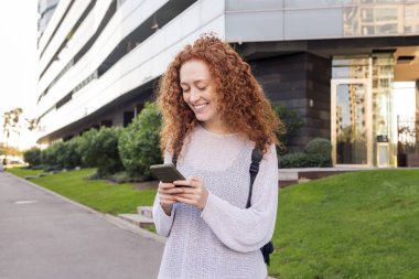 Kıvırcık saçlı genç bir kadın cep telefonuna bakarken mutlu gülümsüyor, iletişim teknolojisi ve modern yaşam tarzı kavramı, metin için metin alanı