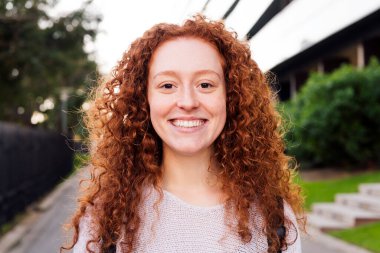 Kıvırcık saçlı genç bir kadının portresi. Kameraya, mutluluk konseptine ve şehir yaşam tarzına bakarken mutlu gülümsüyor.