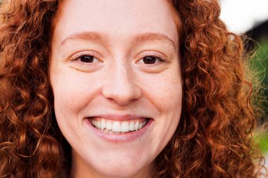 Kameraya, gençlik konseptine ve şehir yaşam tarzına bakarken mutlu gülümseyen kıvırcık saçlı genç bir kadının yüzünü kapatın.