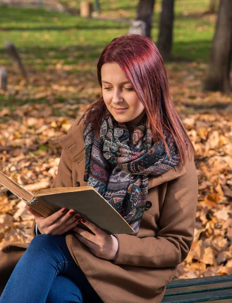 Redhead girl sitting on bench in park and reading book — Stock Photo, Image