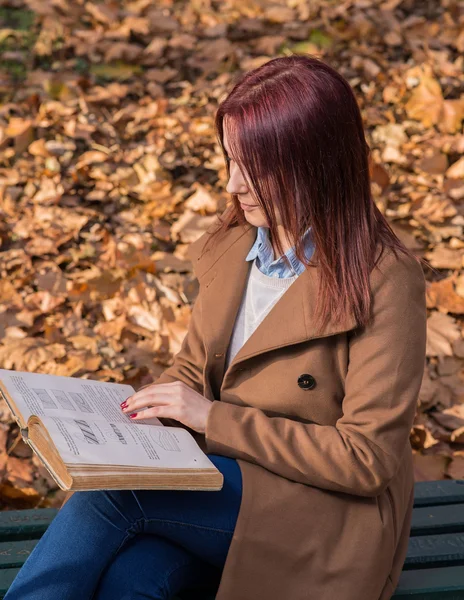 Rödhårig tjej sitter på bänken i park och läsa bok — Stockfoto