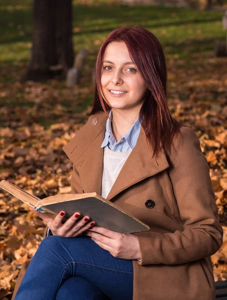 Rödhårig tjej sitter på bänken i park och läsa bok — Stockfoto
