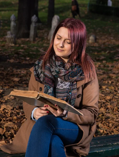 Rödhårig tjej sitter på bänken i park och läsa bok — Stockfoto