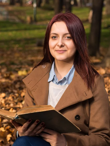 Rödhårig tjej sitter på bänken i park och läsa bok — Stockfoto