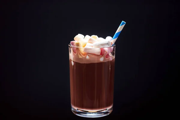 Un vaso de chocolate caliente con malvaviscos de colores y una pajita para beber sobre un fondo negro. — Foto de Stock