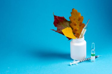 Sonbahar yapraklı beyaz hap kavanozu, civa termometresi, ampel ve şırınga mavi arka planda. Soğuk algınlığı