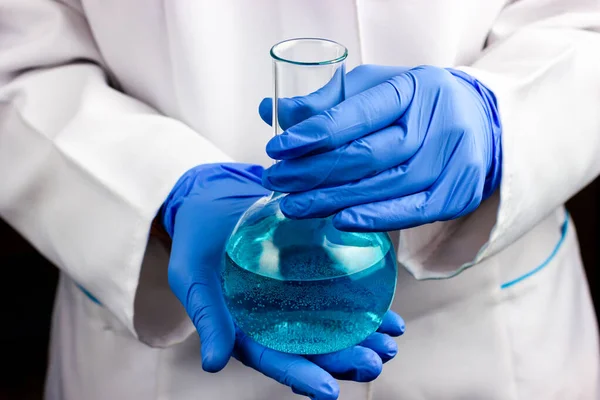 Een chemicus in blauwe handschoenen en een witte chemische vacht heeft een chemische kolf met een blauwe vloeistof. — Stockfoto