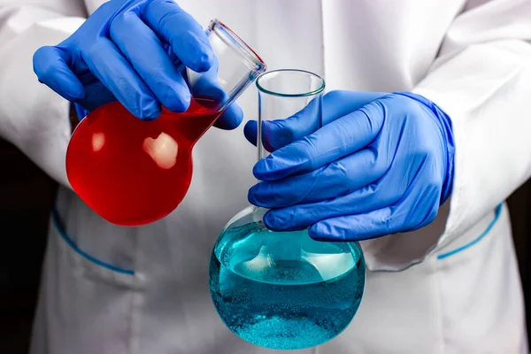 Um químico com um casaco branco e luvas azuis segura dois frascos químicos com líquido — Fotografia de Stock