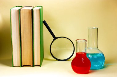 Üç kitap, bir büyüteç ve sarı arka planda kimyasal mataralar. Bilimsel araştırma, çalışma.