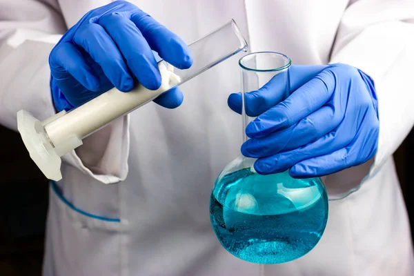 Um químico com um casaco branco e luvas azuis está realizando pesquisas. Interacções com o tubo de ensaio e o balão químico — Fotografia de Stock