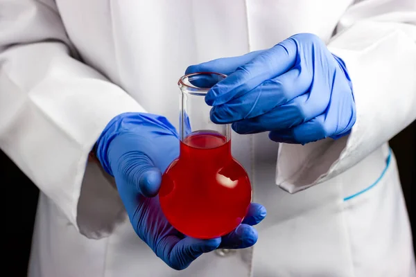 Un chimico con un cappotto bianco e guanti blu tiene una fiaschetta di vetro. Laboratorio chimico, condurre esperimenti chimici. — Foto Stock