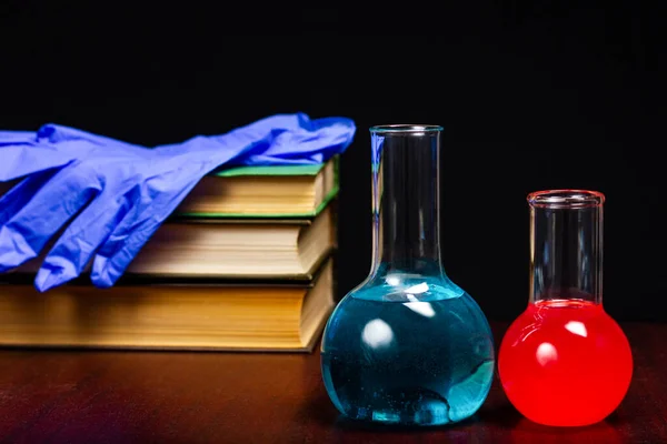 Glass chemical flasks with liquid and several textbooks on a dark wooden table. Class in chemistry, school, student study of science.