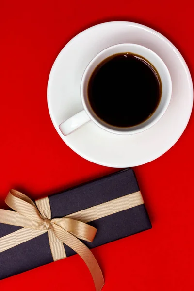 Šálek kávy a černý dárek s béžovou stuhou na červeném pozadí. Pohled shora. — Stock fotografie